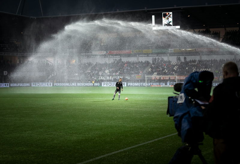 footballer warms up with sprinkler watering pitch in foreground at stadium